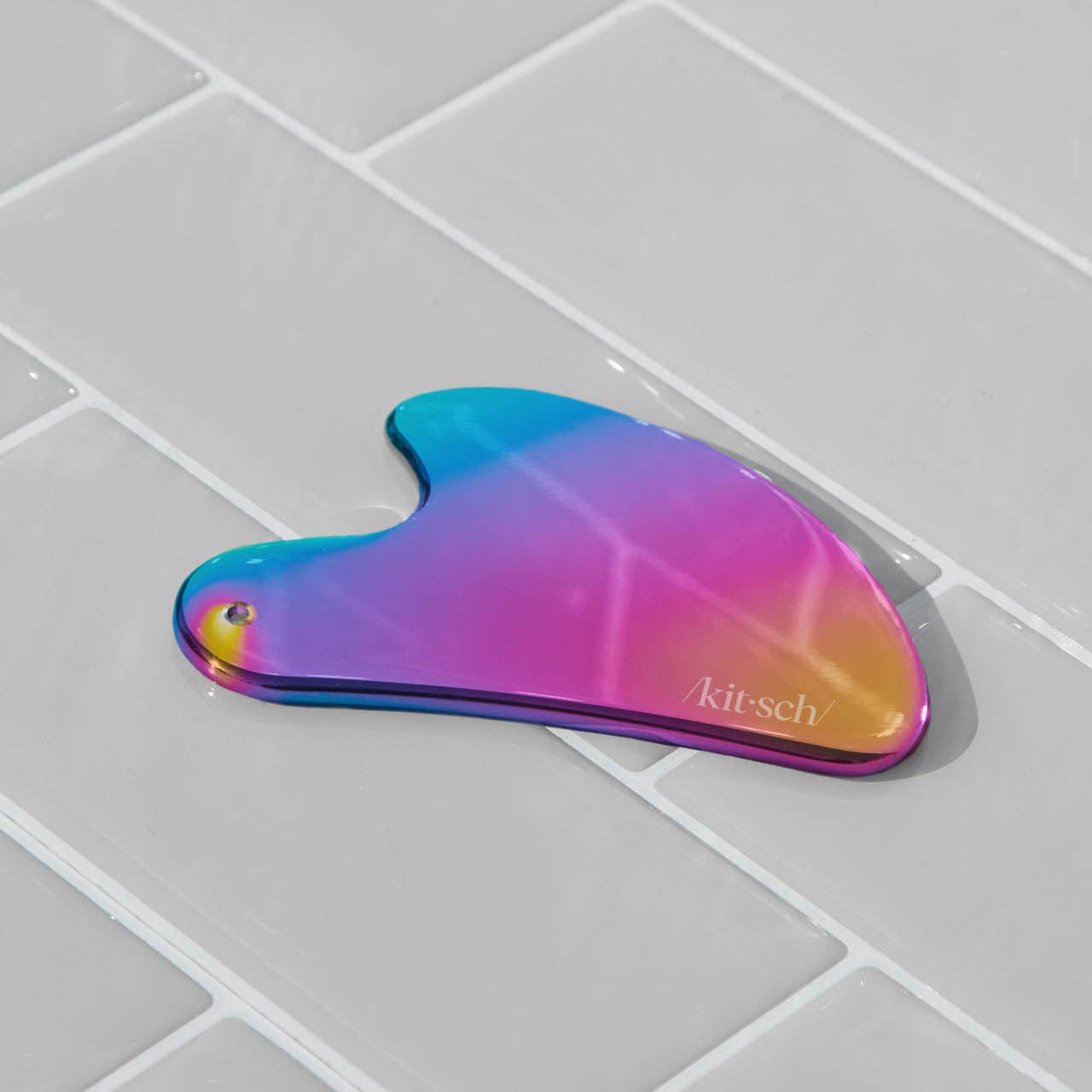 Kitsch Stainless Steel Gua Sha - Iridescent on light gray tiles, highlighting its smooth, curved design for skincare benefits like lymphatic drainage and facial tension relief.