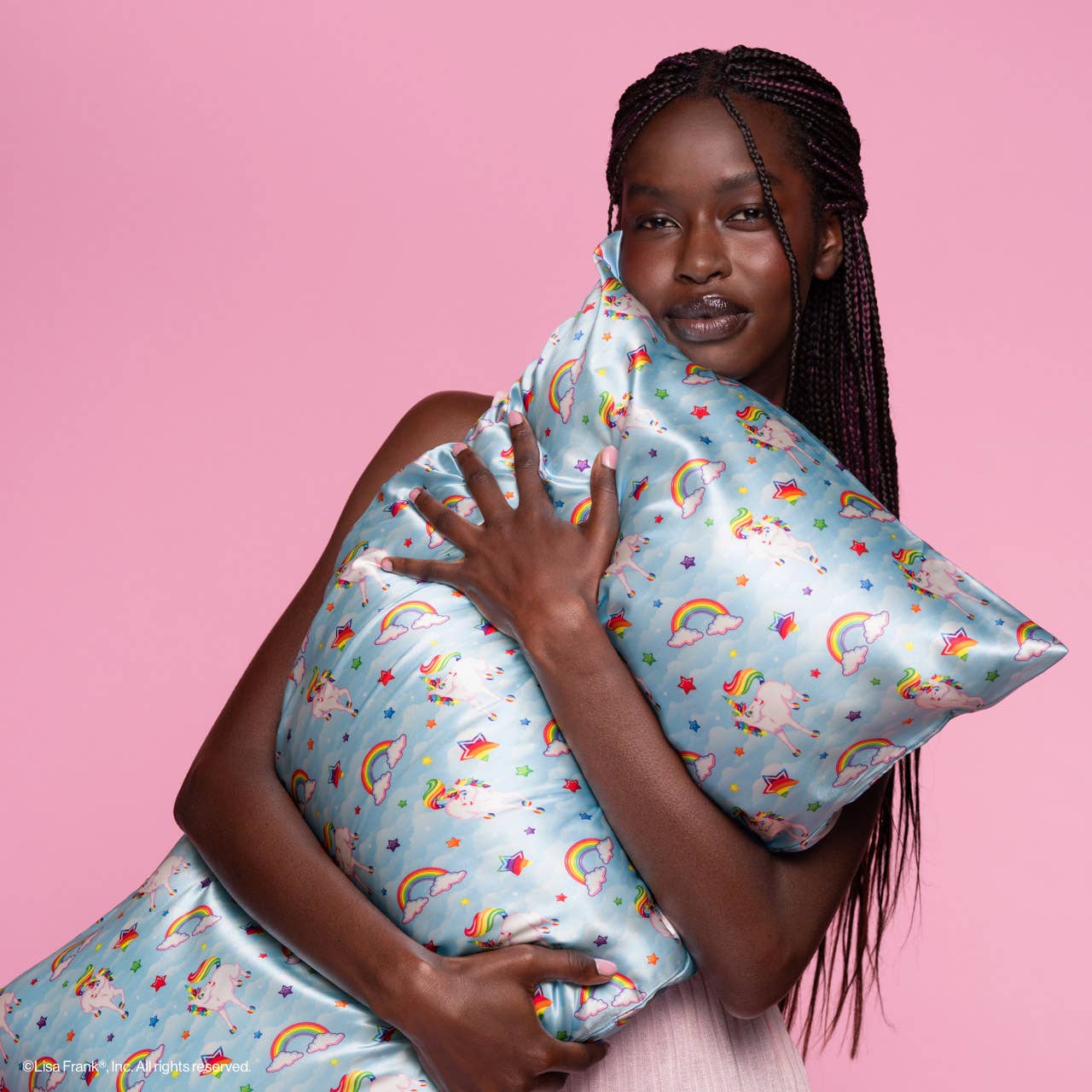 Young Black woman hugs a Lisa Frank x Kitsch Standard Satin Pillowcase featuring unicorns, rainbows, stars; designed to reduce frizz, breakouts, and facial creases.