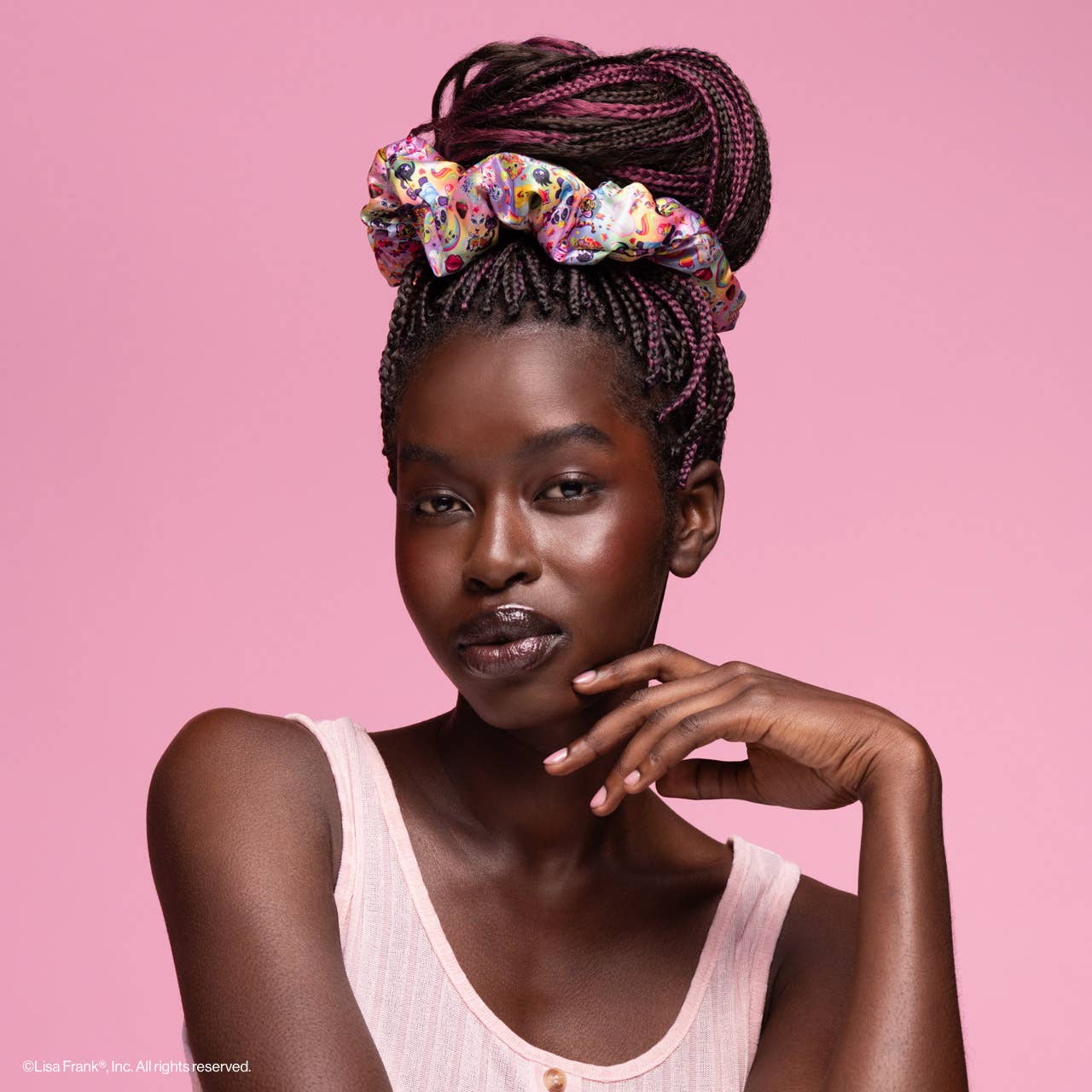 Young Black woman with smooth complexion wears Lisa Frank x Kitsch Pillow Scrunchies® in Satin, showcasing colorful motifs atop her high bun against a pastel-pink background.