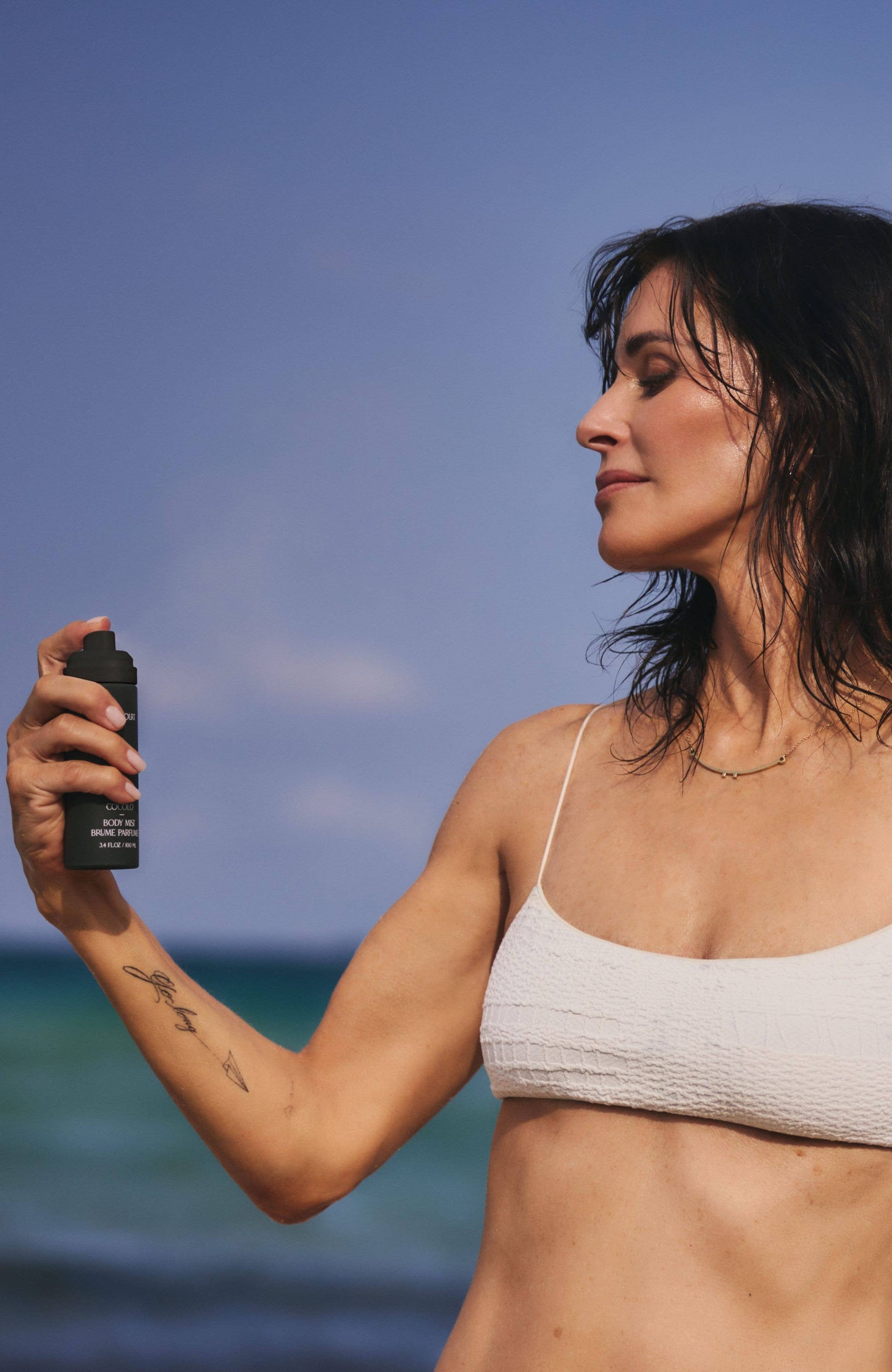 Woman on beach misting herself with Cocomoi Body Mist | Half Case; features a luxurious, eco-friendly fragrance, enhancing personal scent with coconut, cedarwood, and fig notes.