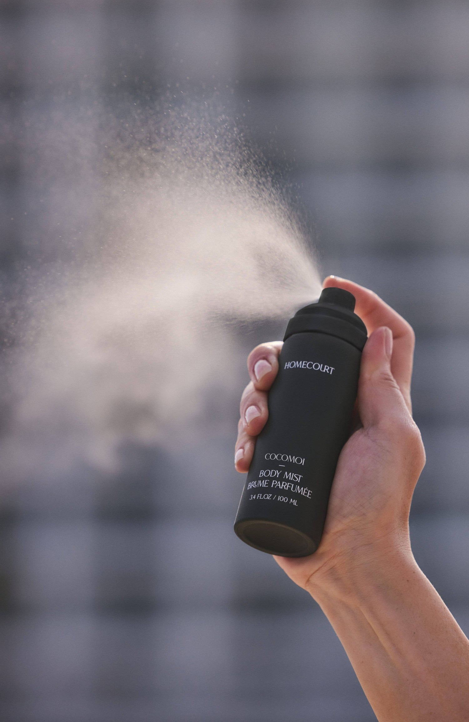 Hand holding a matte-black Cocomoi Body Mist spray bottle, releasing a fine mist, highlighting its eco-friendly, sustainable fragrance by Homecourt.