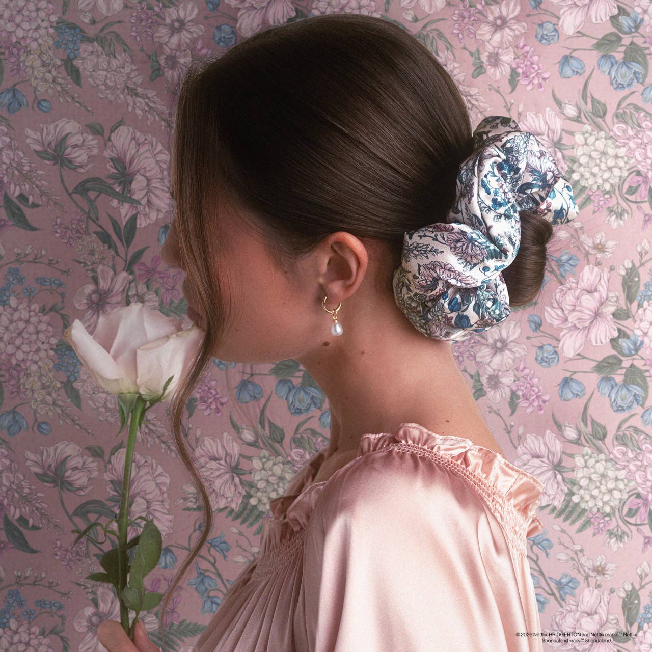 Young woman with dark hair in a low bun, wearing a Bridgerton x Kitsch Satin Pillow Scrunchie® in Toile in Bloom, gently smelling a pale rose.