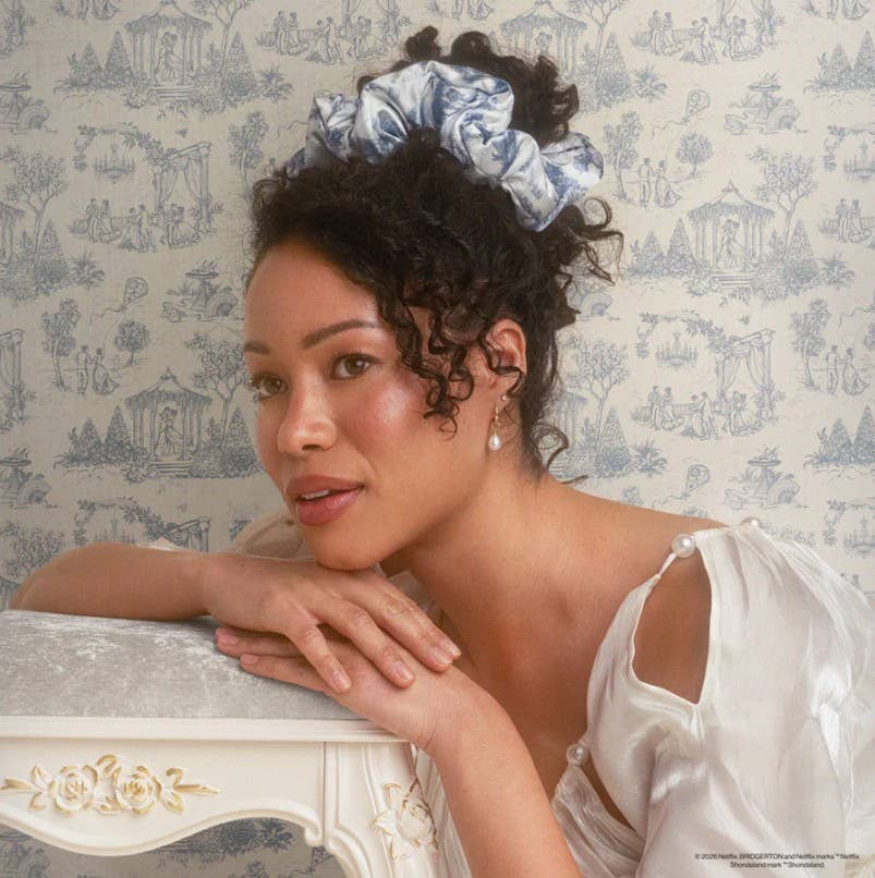 Woman with loose curls wears Bridgerton x Kitsch Satin Pillow Scrunchie® in Toile in Bloom, leaning on ornate table against toile wallpaper.
