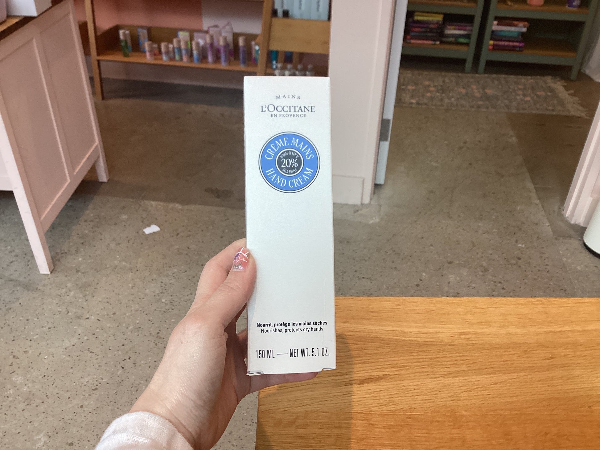 Close-up of a hand holding a box of L’Occitane Hand Cream with 20% shea butter, 150 ml, amidst beauty products on store shelves.