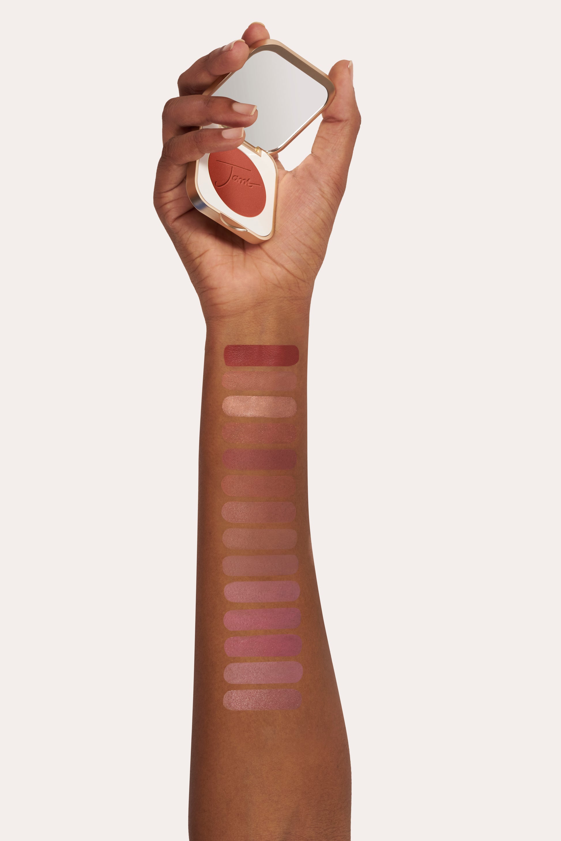 Dark-skinned forearm displays PurePressed® Blush swatches; hand holds compact with reddish-orange powder, showcasing the buildable, silky mineral formula for a natural flush.