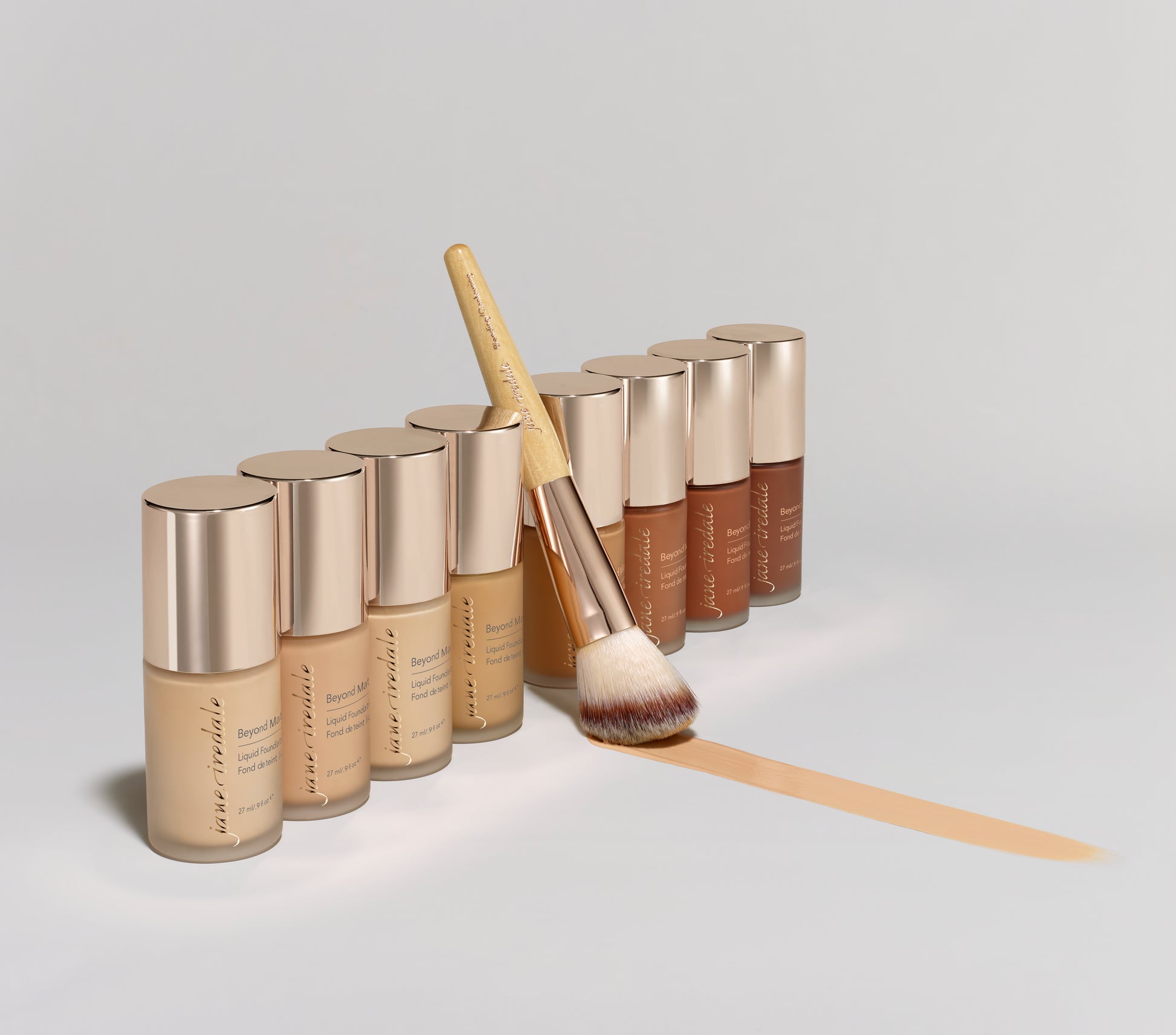 Row of jane iredale Beyond Matte Liquid Foundation bottles in shades, with a makeup brush; a swatch showcases buildable, semi-matte, plant-based formula.