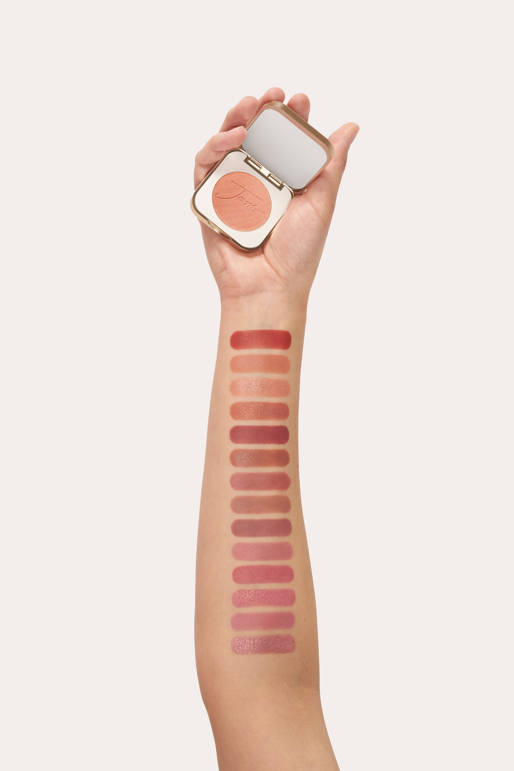Hand holding PurePressed® Blush compact with peachy-coral shade; forearm displays 15 swatches from coral to pink, showcasing the product's versatile, buildable mineral formula.