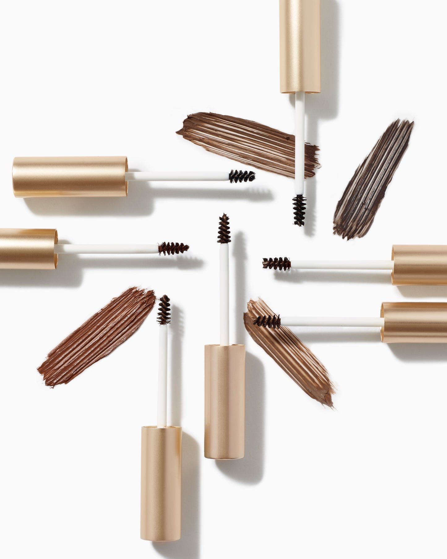 PureBrow® Brow Gel tubes with spoolie brushes arranged radially; swatches show brown tints. Defines, grooms, and sets brows with a natural finish.