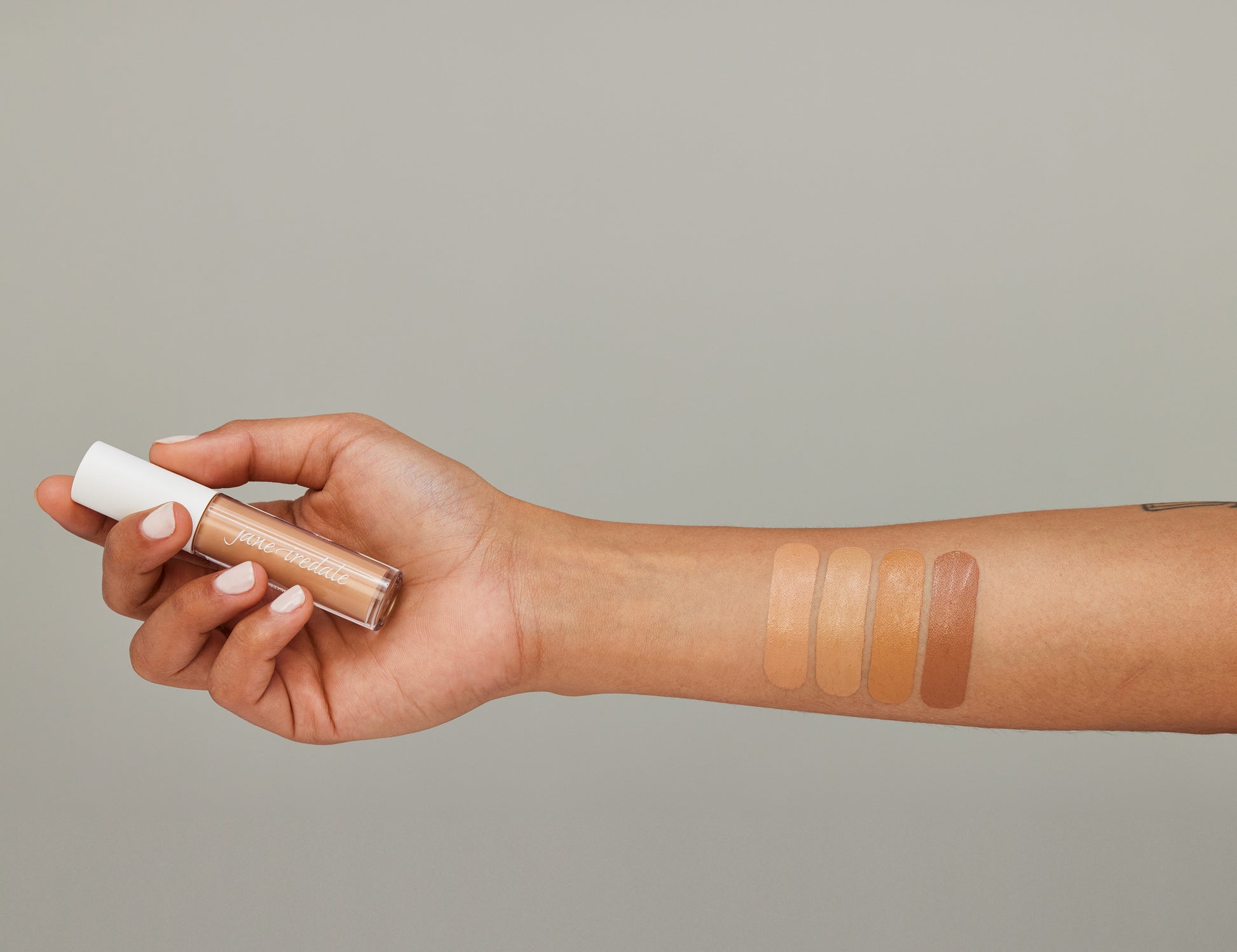 Forearm with four swatches of PureMatch™ Liquid Concealer by Jane Iredale, showcasing shade range and a hand holding the product tube.