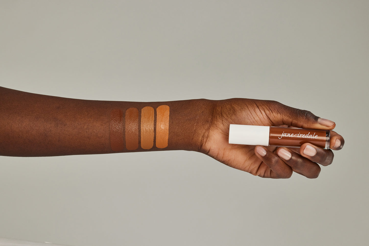 Forearm displaying four liquid swatches with a hand holding a jane iredale PureMatch™ Liquid Concealer tube, demonstrating shade range and natural coverage.