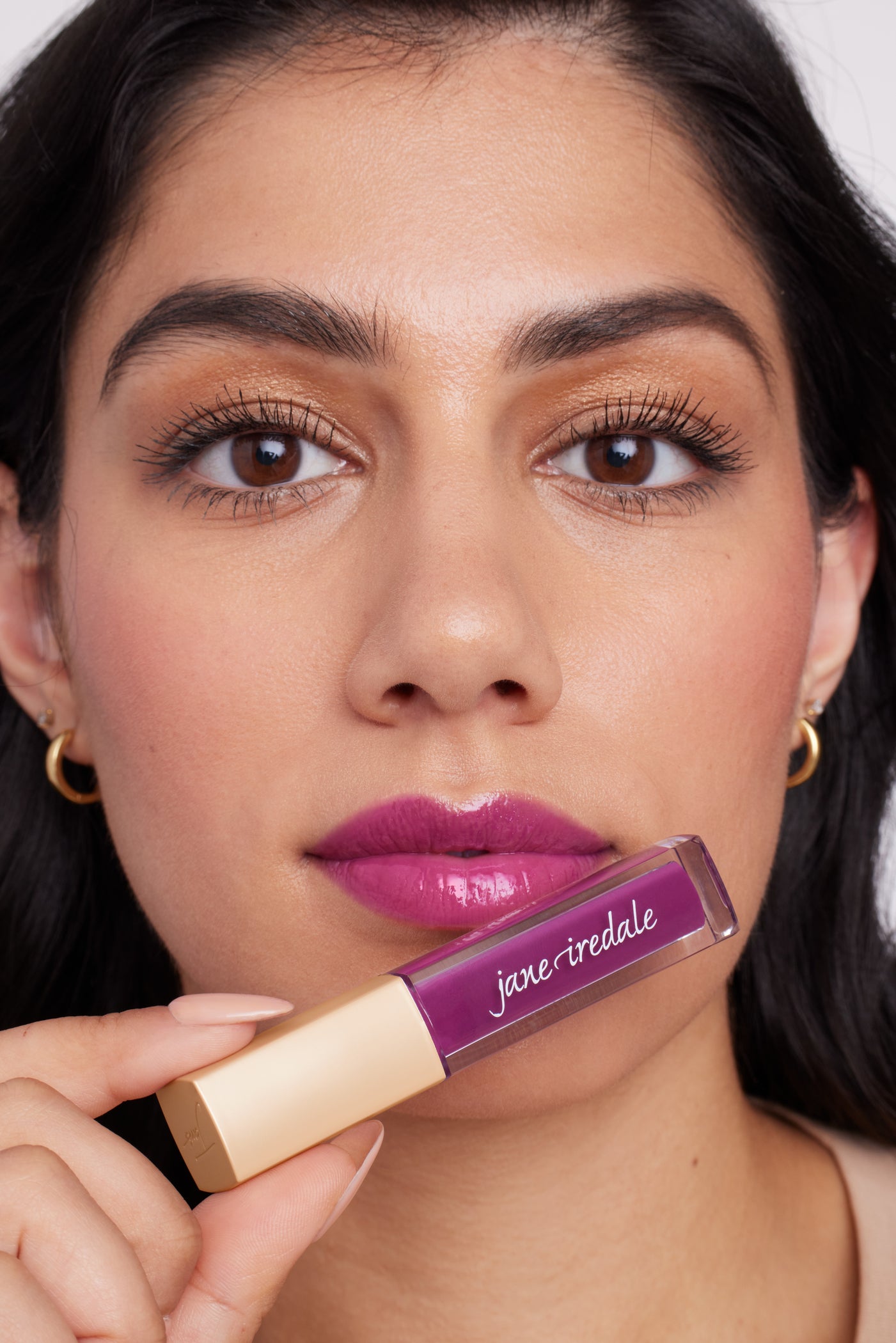 Woman wearing glossy purple ColorLuxe High Impact Lip Glaze by jane iredale, showcasing its bold, glossy finish and high pigmentation, holding matching tube.