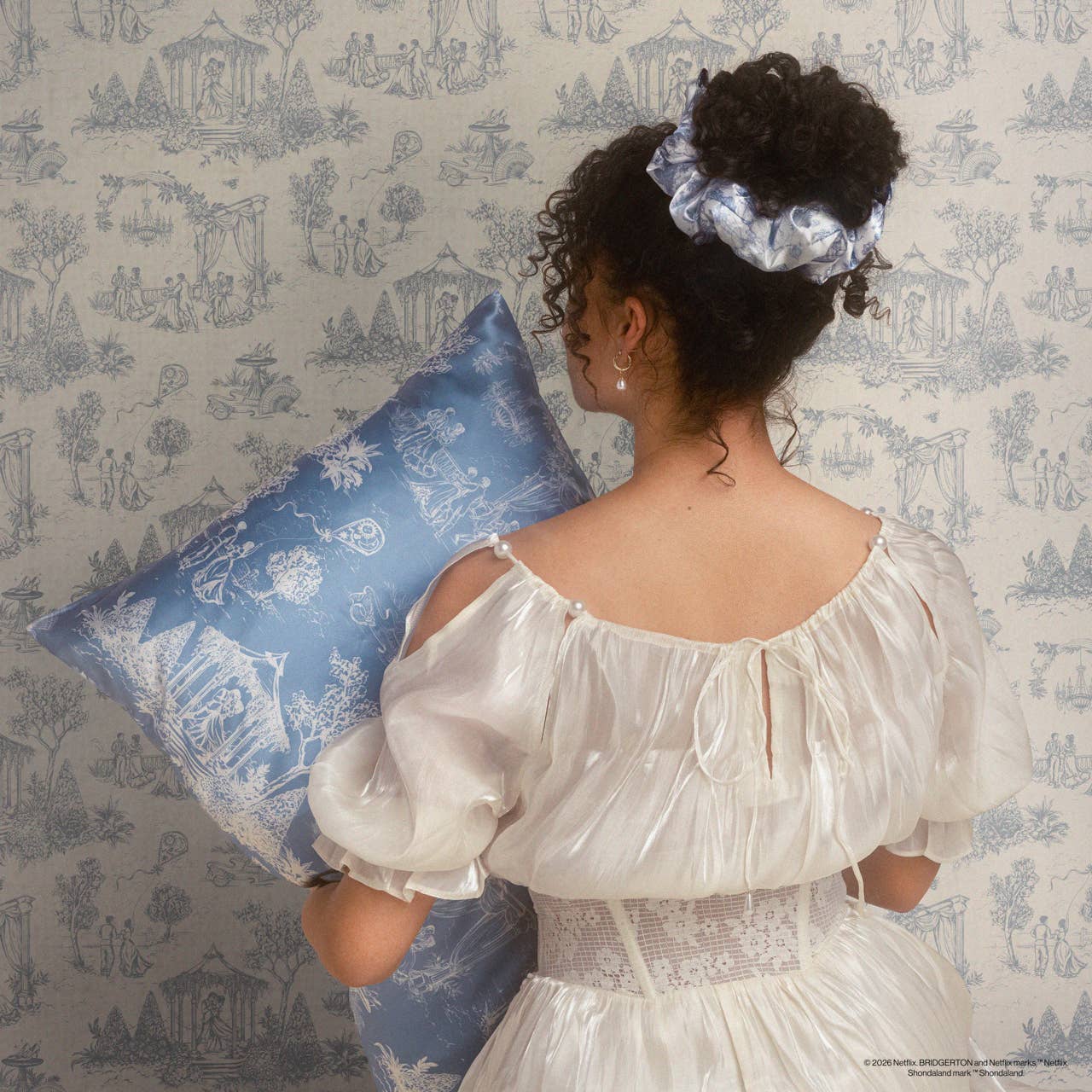 Woman in Regency-style gown holding a decorative cushion, wearing a Bridgerton x Kitsch Satin Pillow Scrunchie® in Toile in Bloom, matching the cushion and wallpaper.