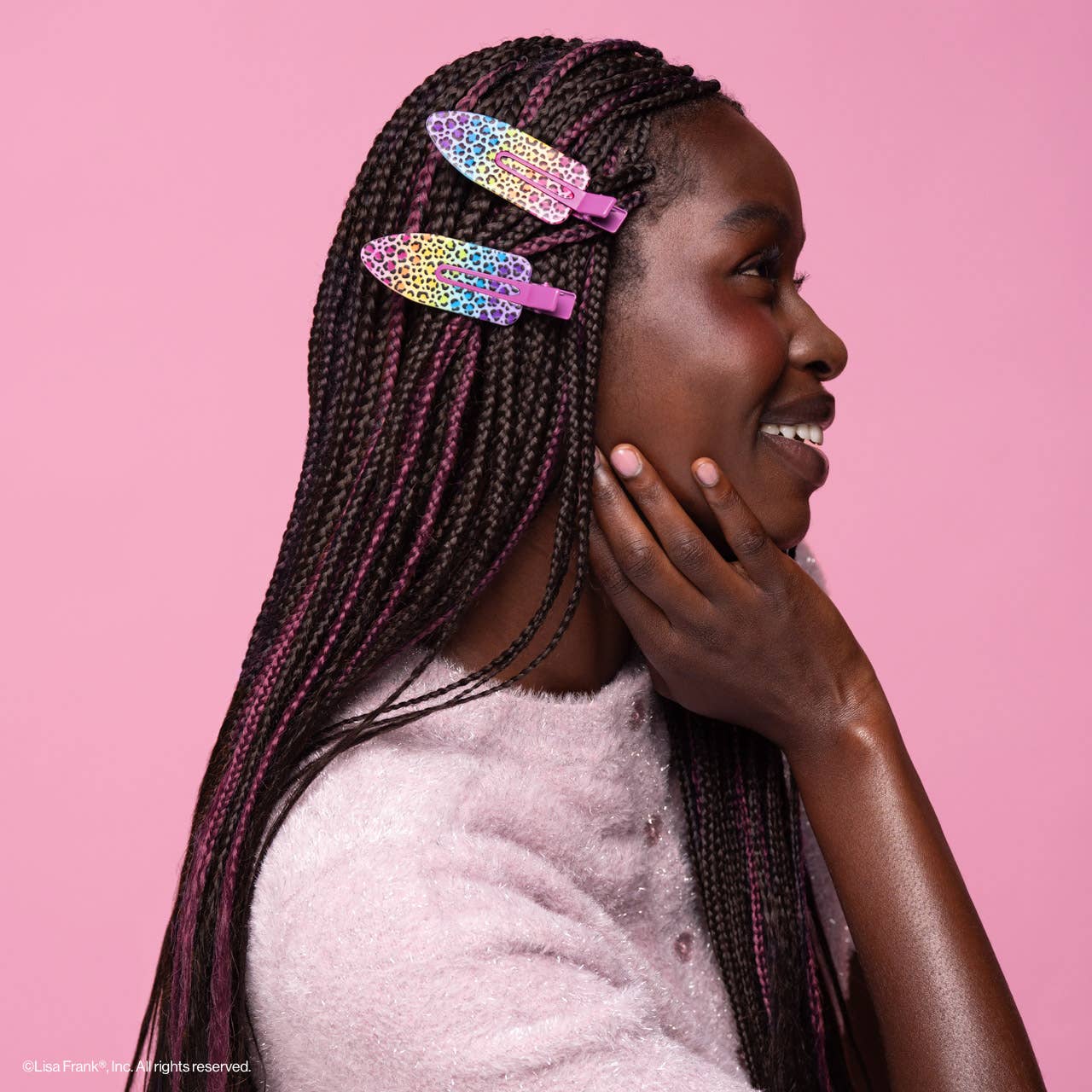 Young person with box braids wears Lisa Frank x Kitsch XL Creaseless Clips in Rainbow Leopard, pinning back hair, smiling in a light pink sweater.