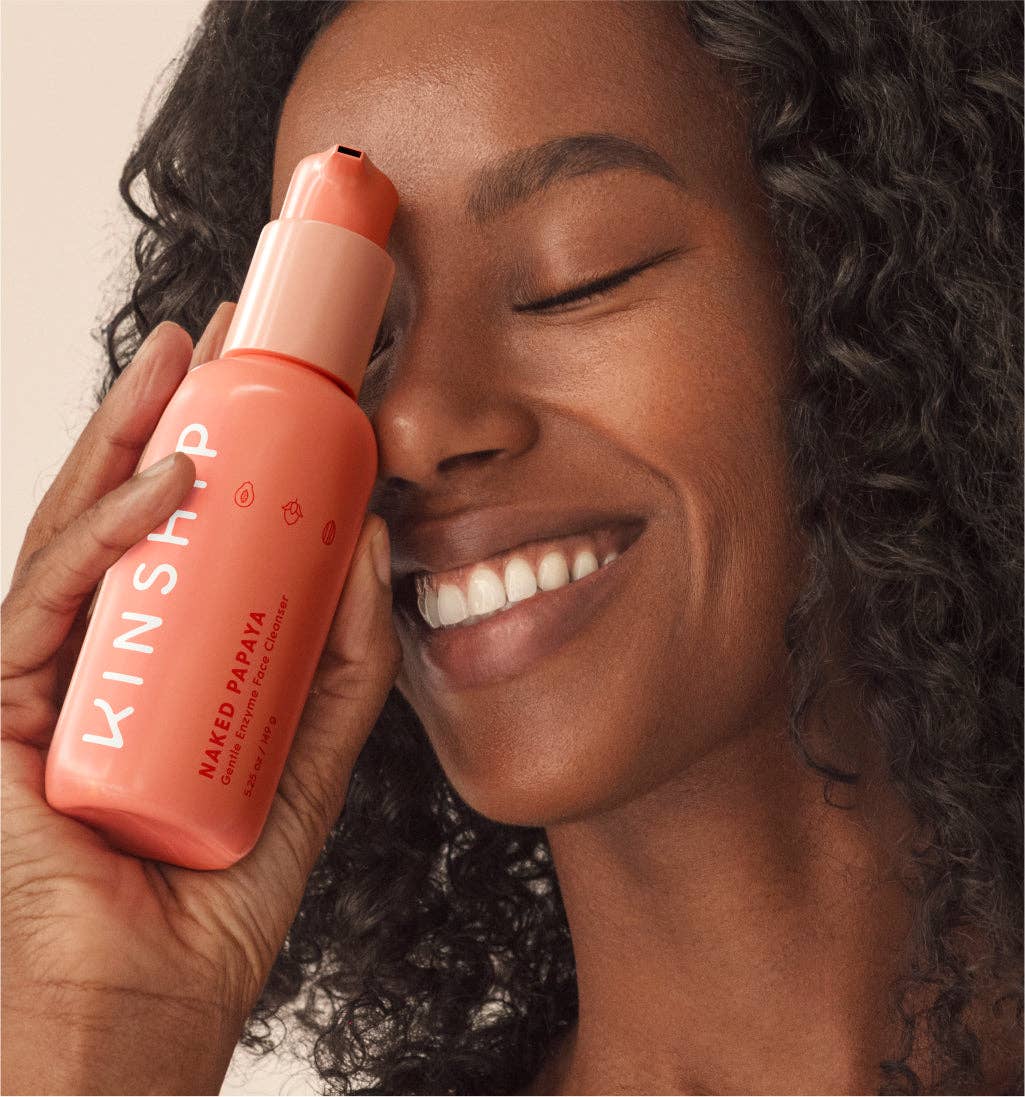 Woman smiling, holding KINSHIP Naked Papaya Gentle Enzyme Milky Cleanser bottle, highlighting its award-winning, dermatologist-recommended formula for hydrating and cleansing sensitive skin.