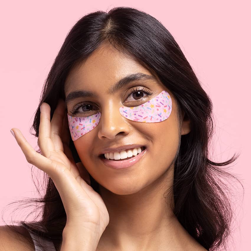 Young woman wearing Eye Want Cake Smoothing & Hydrating Eye Patches, showcasing smooth skin and vibrant under-eye area with sprinkle design against a soft pink background.