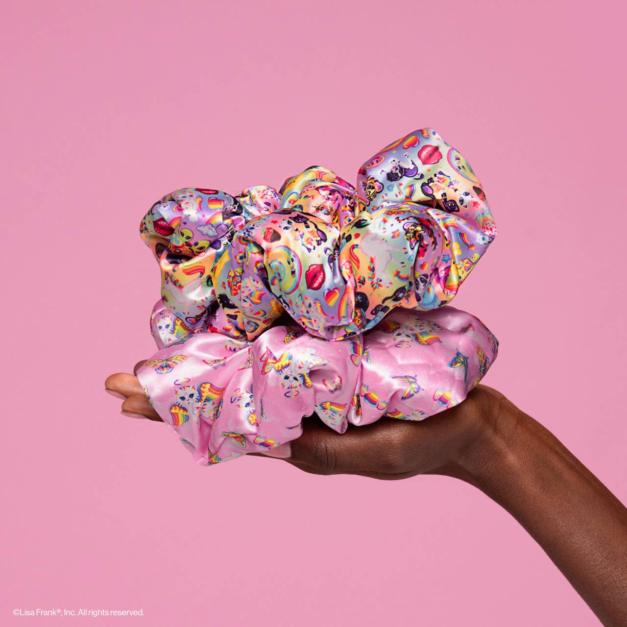 Hand holding two oversized satin scrunchies, Lisa Frank x Kitsch Pillow Scrunchies® in Satin, featuring unicorn prints and a colorful collage, against a pink backdrop.