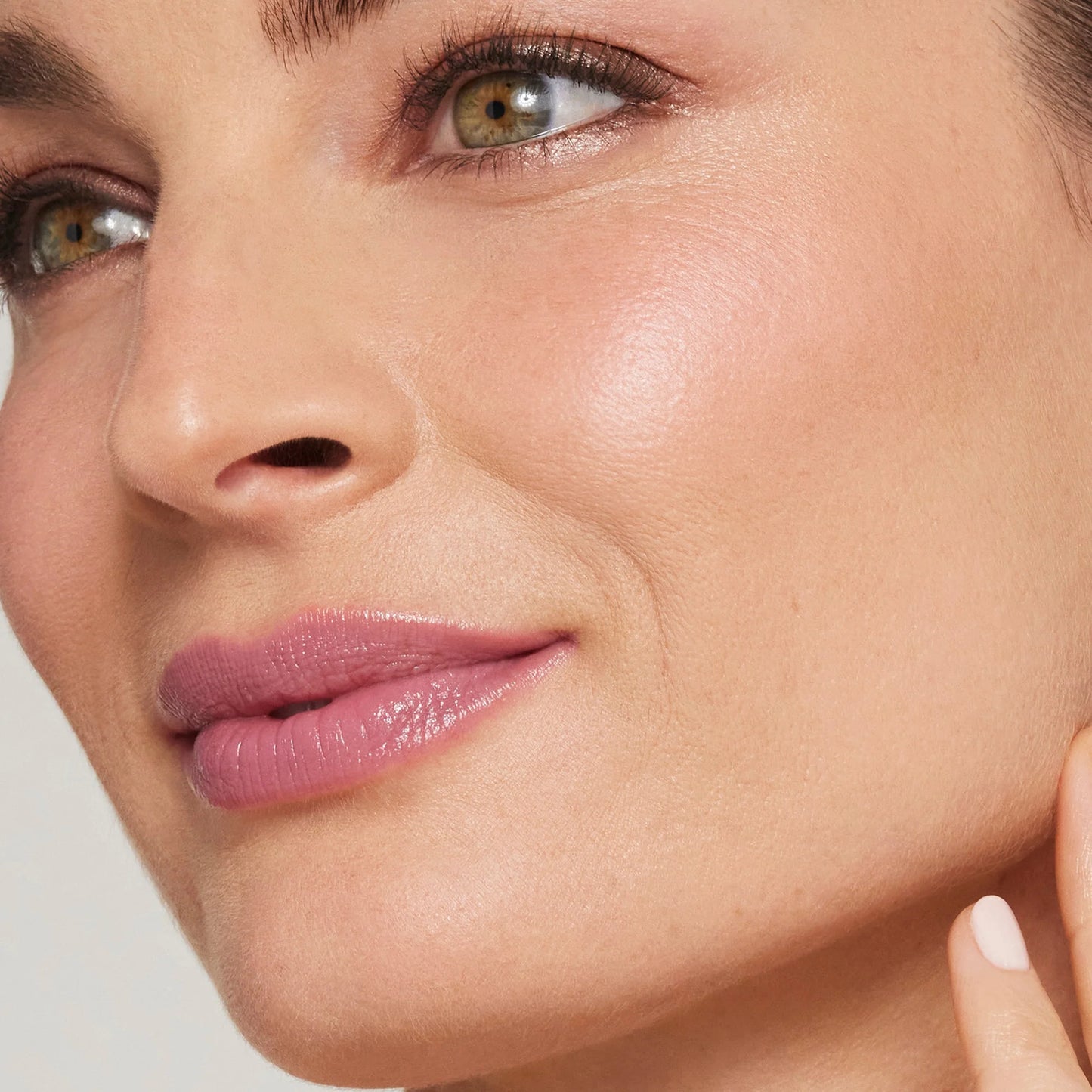 Woman with pinkish-mauve lips and subtle eye makeup, showcasing the Glow Time® Blush Stick's natural finish on smooth cheeks, touching jawline with manicured fingernail.