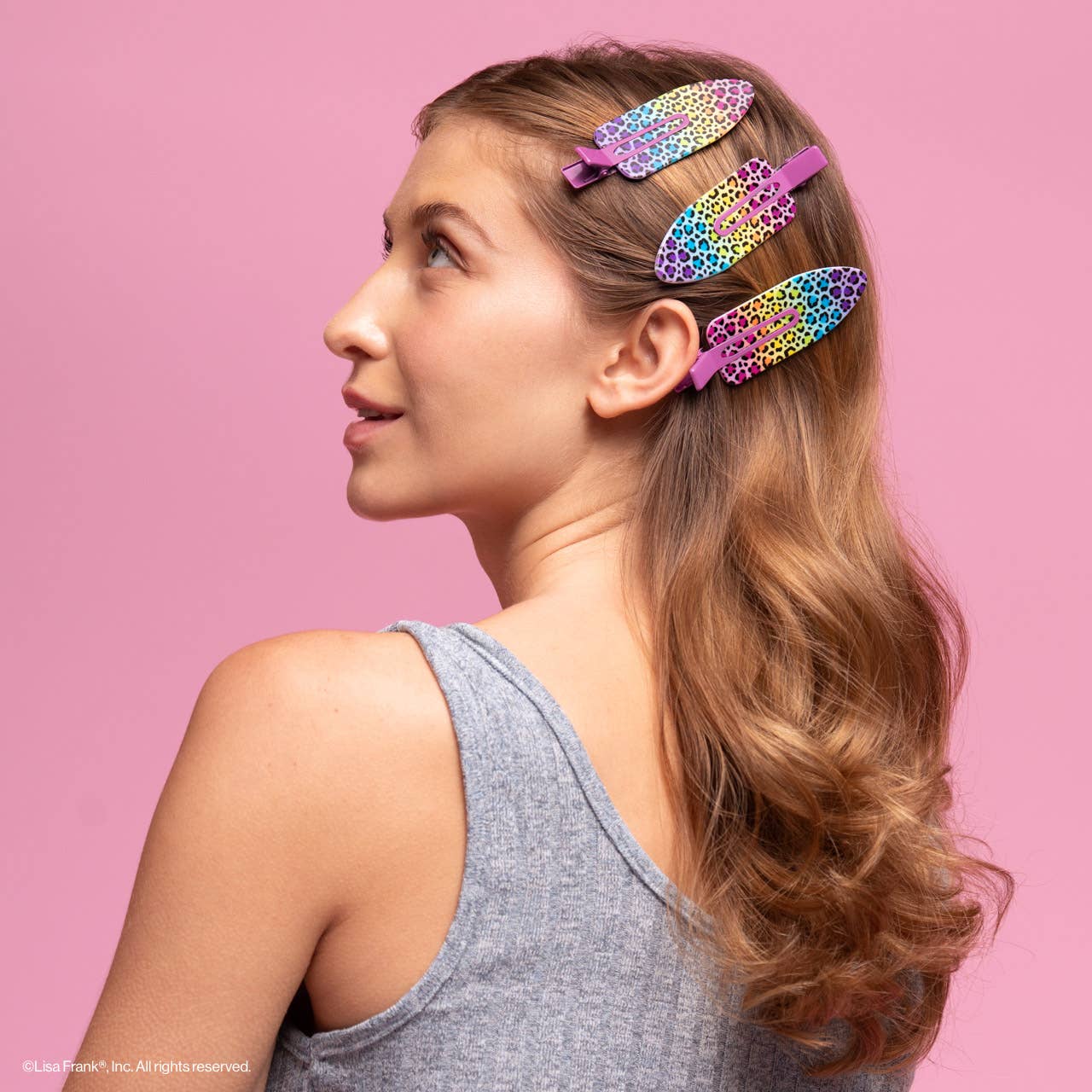 Young woman wearing Lisa Frank x Kitsch XL Creaseless Clips in Rainbow Leopard, securing hair with vibrant, oversized clips against a pink backdrop.