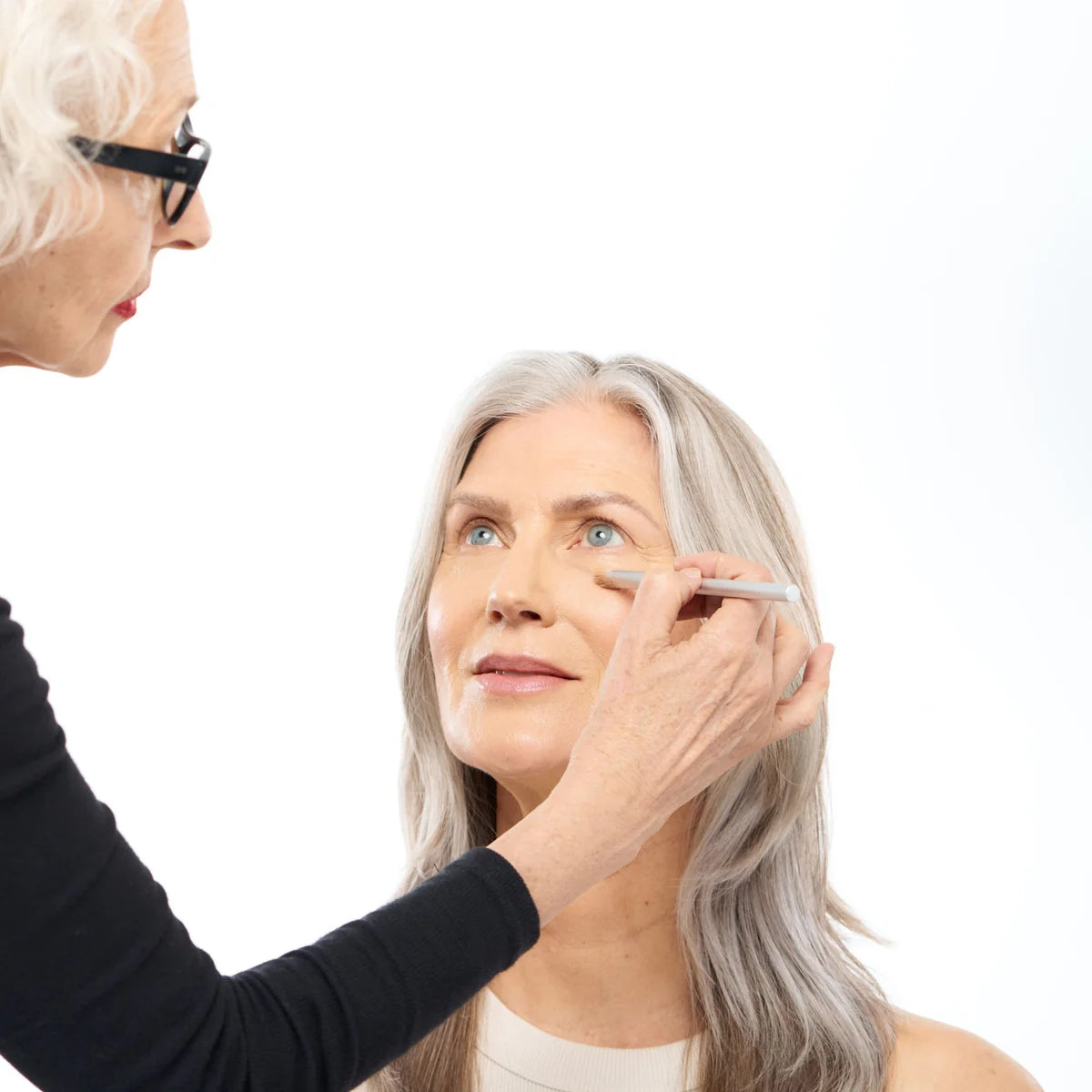 A makeup artist uses the Skin2Skin Concealer Brush with soft vegan bristles to apply product under a mature woman's eye, showcasing precision and gentle application.